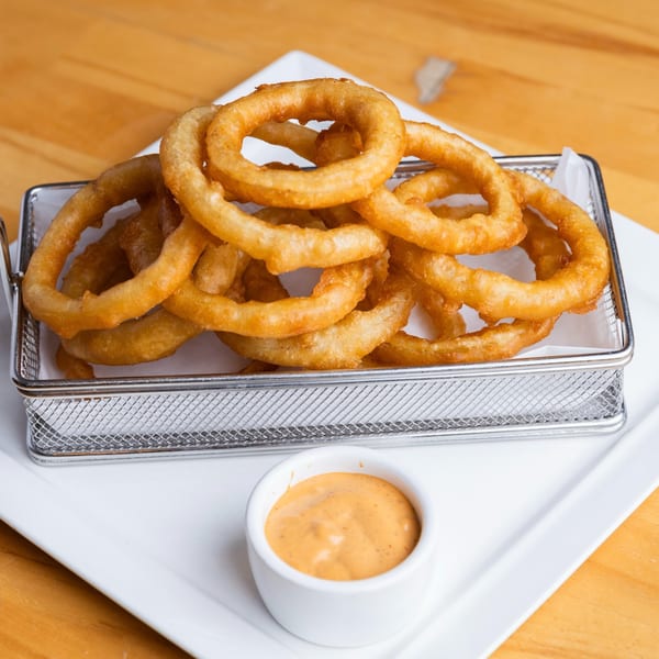 Golden-brown onion rings in a metal basket with a side of dipping sauce on a white plate.